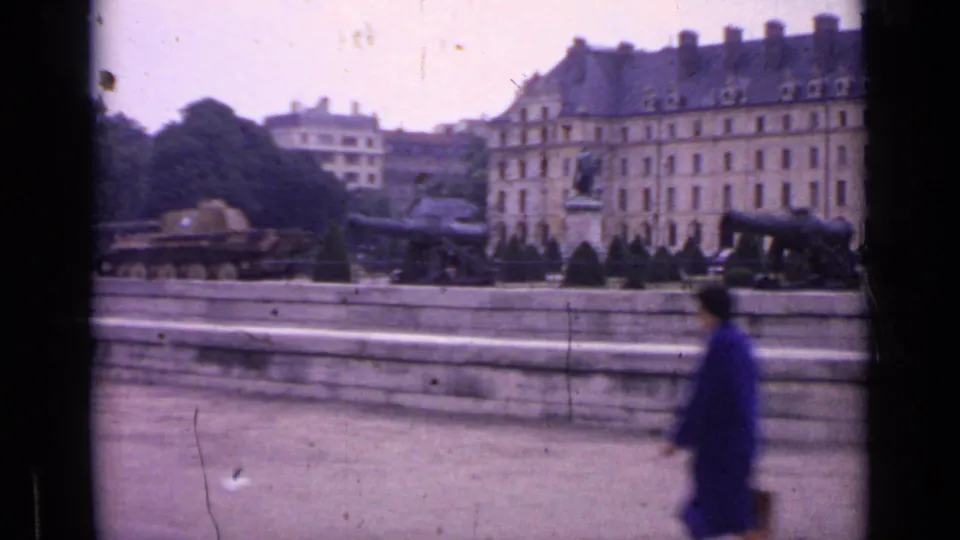1962 Paris home-movie frame of a long building, formal garden, cannons, tank, statue, and blurred pedestrian near a stone embankment.