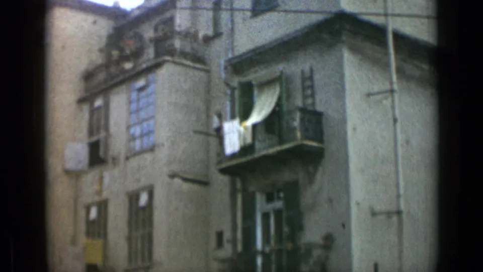 1962 Italy-labeled 8mm frame of a weathered apartment facade with balcony laundry, shutters, windows, and small plants.