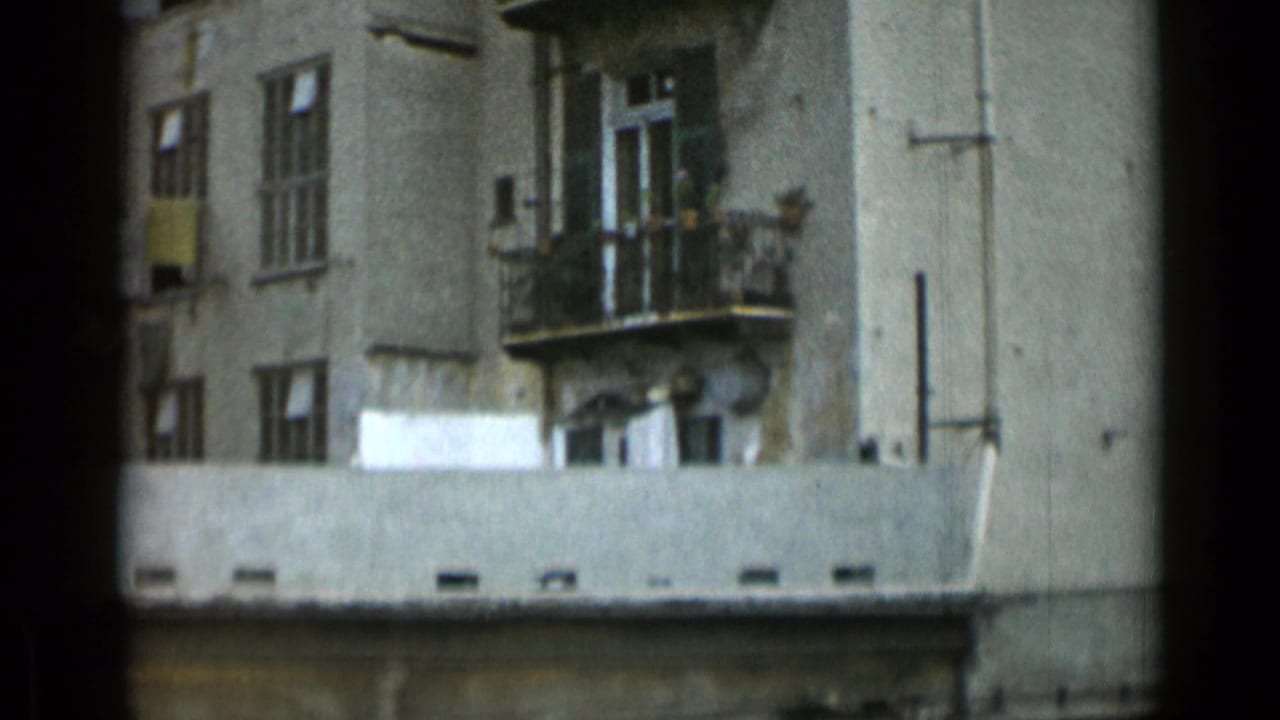 Apartment building corner with balconies, windows, rooftop edge, and exterior pipes.
