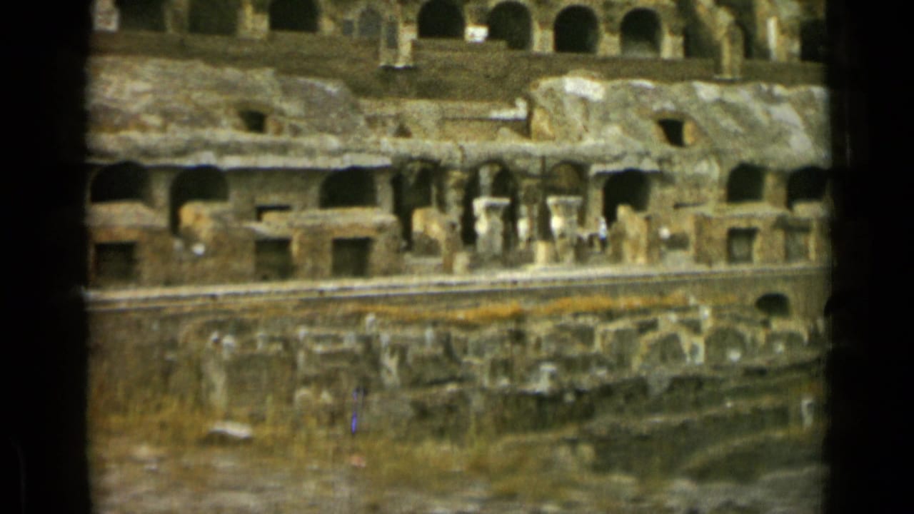 Interior Colosseum ruins with arched stone walls, openings, and small distant visitors.
