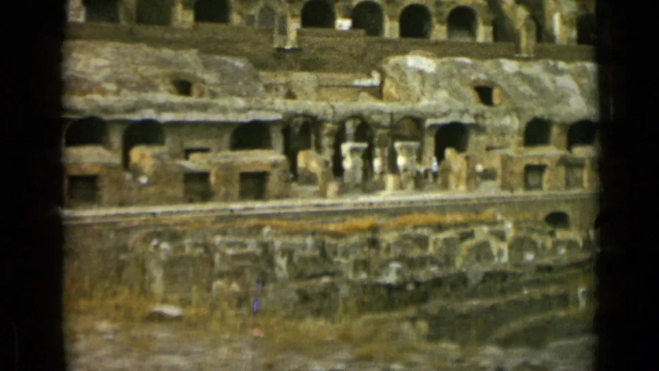 1962 8mm view inside Rome's Colosseum, showing layered stone arches, eroded walls, vegetation, and tiny visitors near the lower level.
