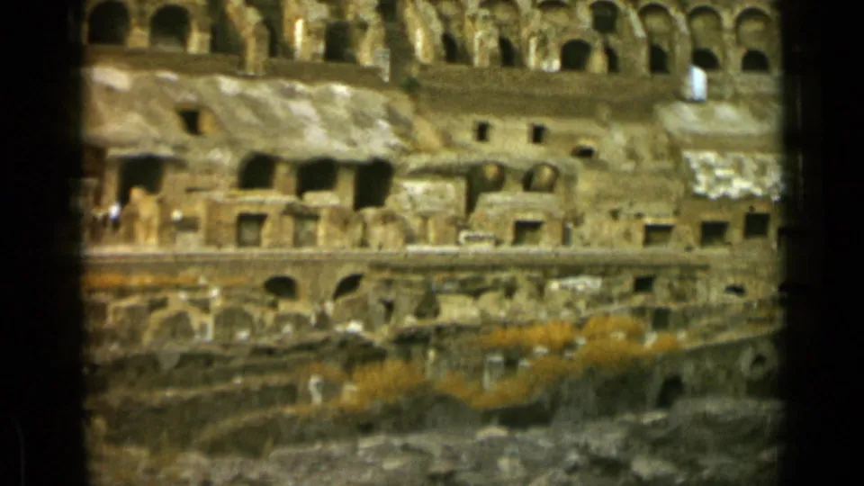 Closer 8mm frame of Rome's Colosseum interior, showing rough stone openings, arched upper corridors, rubble below, and scrub growth along the ruins.