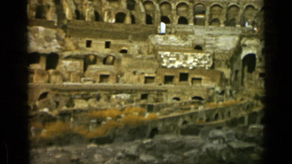 Final 1962 frame inside the Colosseum, with arched galleries above, eroded stone blocks, dark openings, and vegetation across the lower ruins.