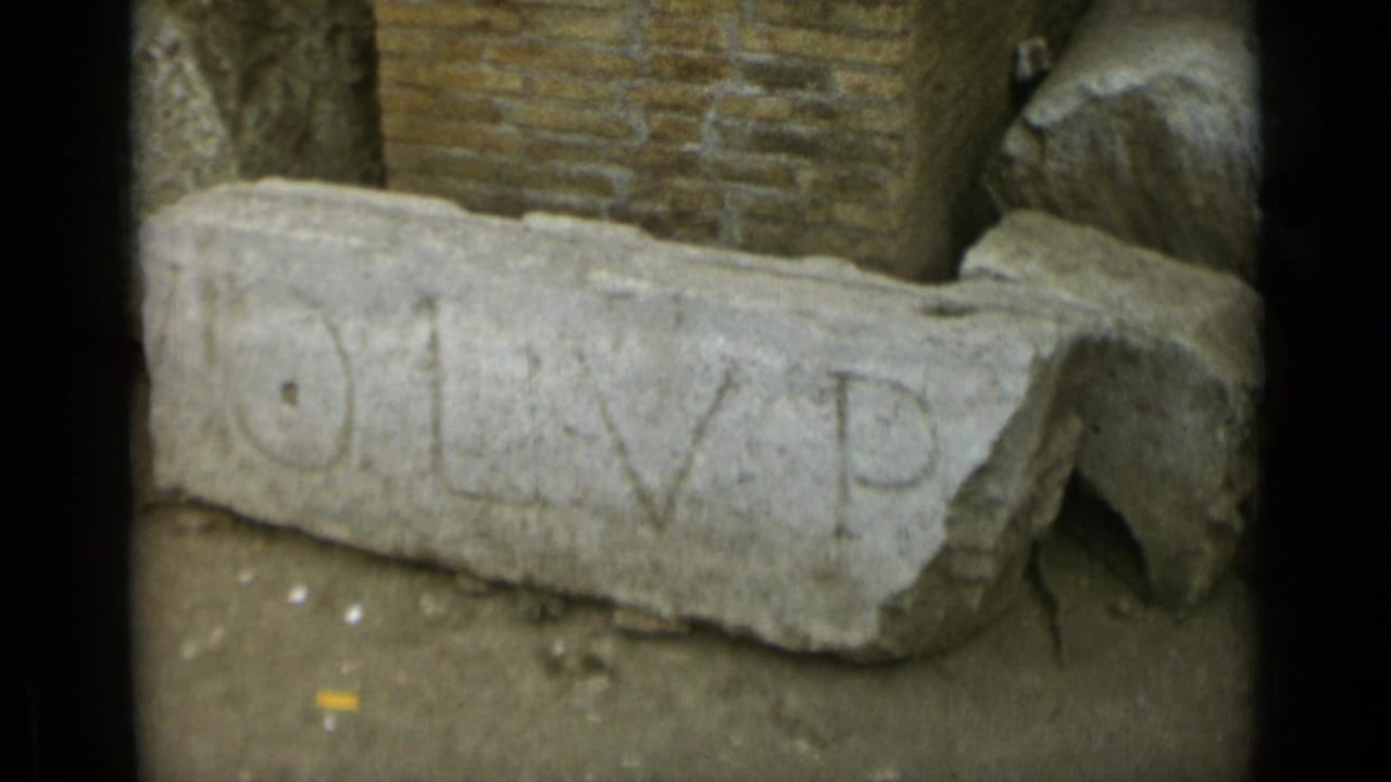 1962 Rome 8mm frame showing a weathered carved stone fragment with partial inscription near ancient brickwork.
