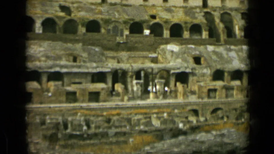 Wider 1962 home-movie view of the Colosseum in Rome, showing layered ruins, arches, railings, and distant visitors.
