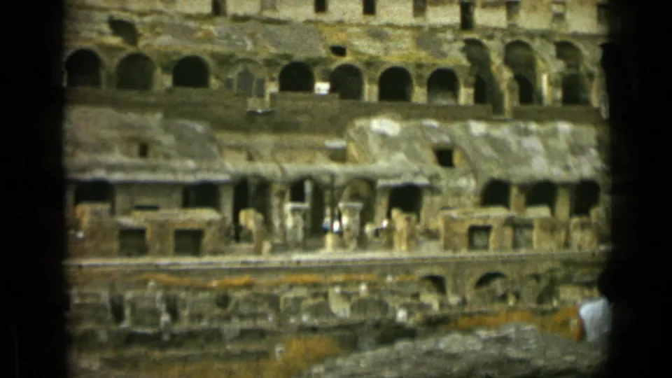 1962 Rome 8mm frame of the Colosseum interior, with repeated arches, rough stone surfaces, and a visitor partly visible near the right edge.