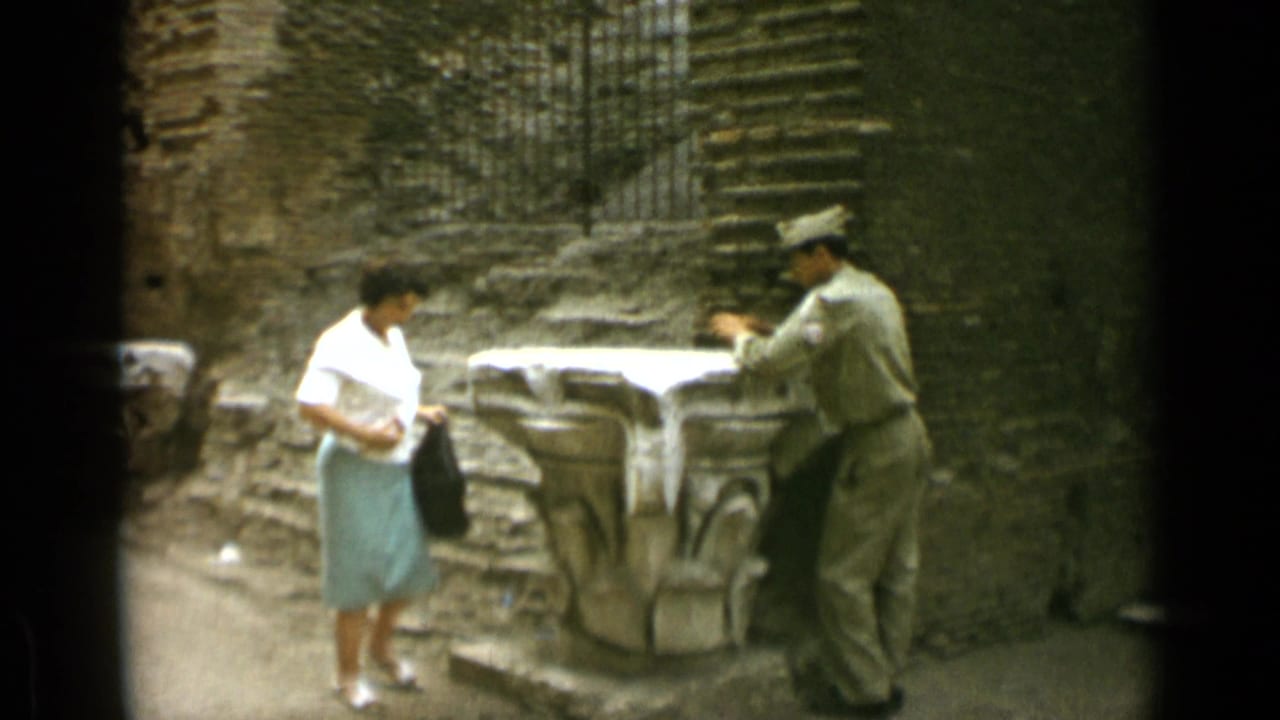 Woman in a white shirt and green skirt stands near a uniformed man by a stone pedestal.