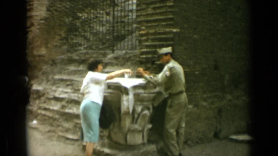A handheld 8mm frame shows the woman leaning toward the uniformed man across the stone pedestal at the Colosseum.