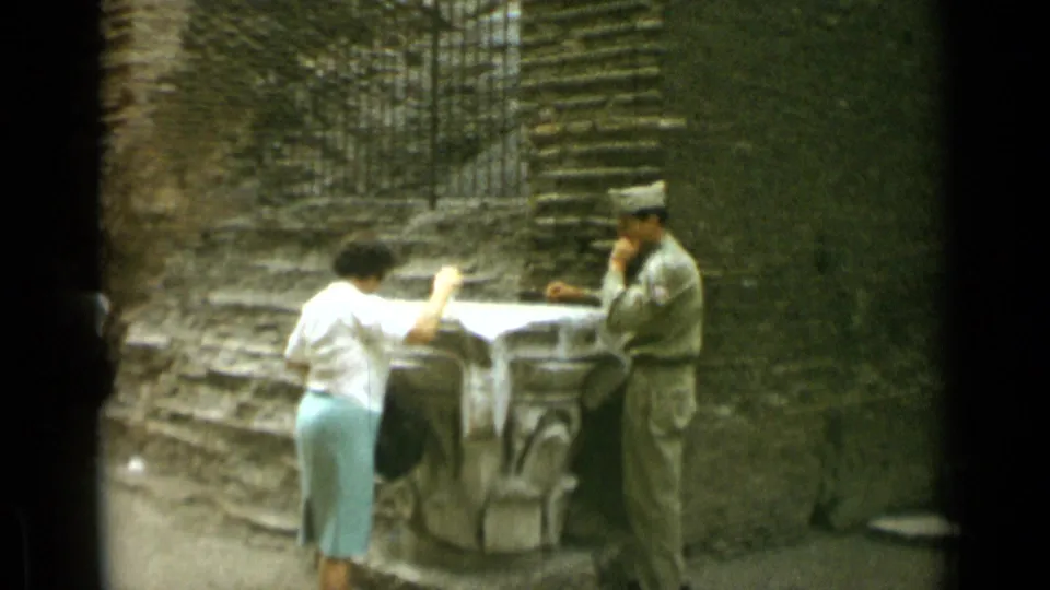 Near the end of the clip, the woman remains by the Colosseum pedestal while the uniformed man stands opposite with his hand near his face.