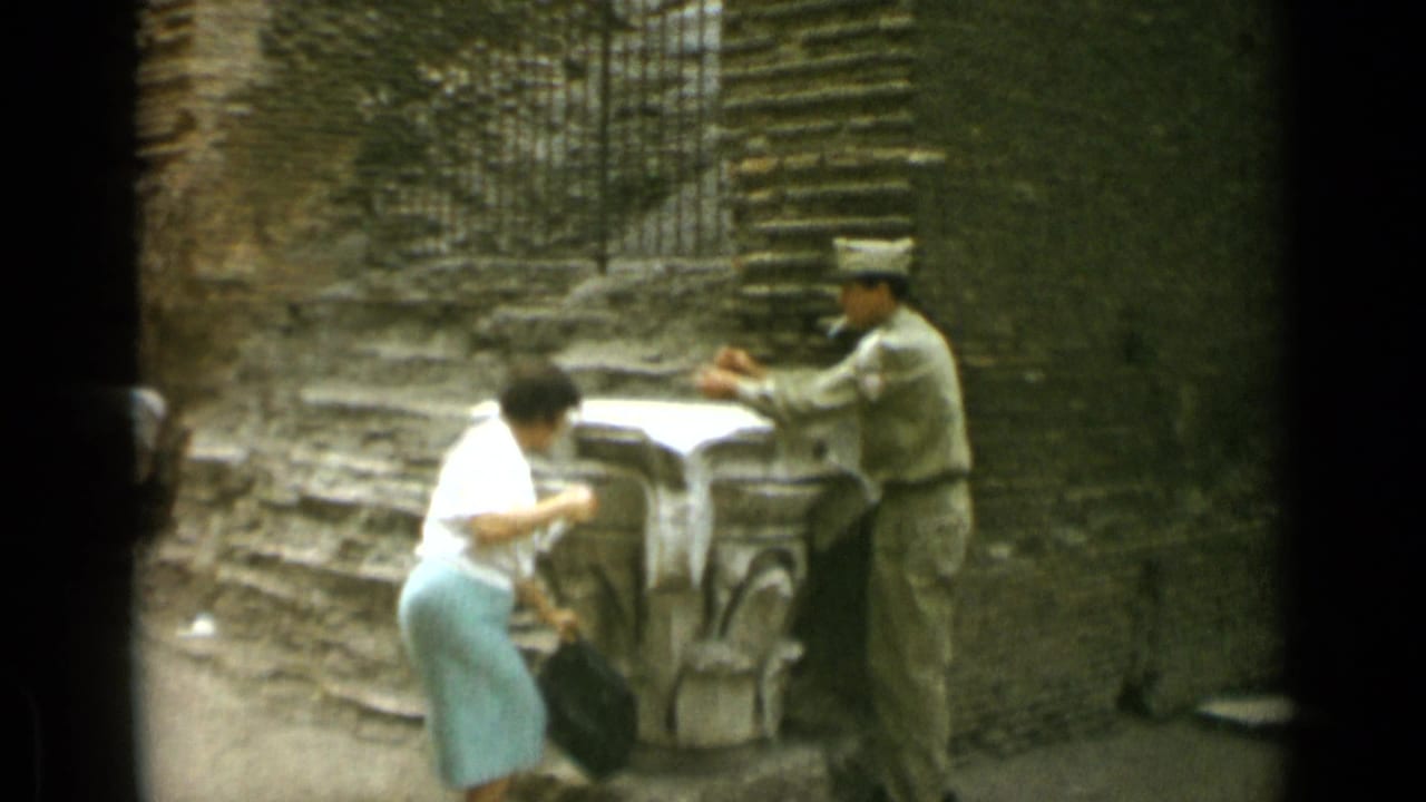 Woman in a white blouse bends near a stone feature while a uniformed man stands opposite.