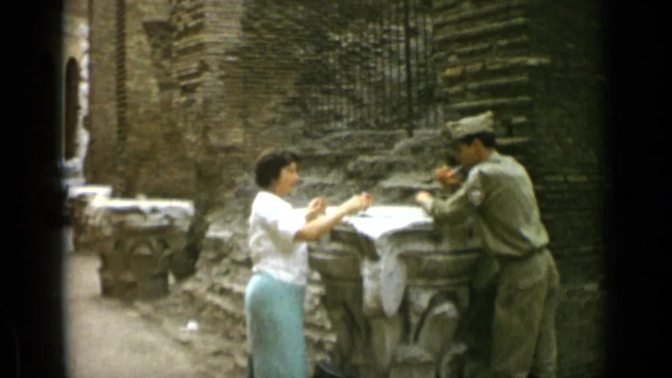 A woman in a pale skirt stands near a uniformed man who appears to smoke beside a stone feature in 1962 Rome footage.