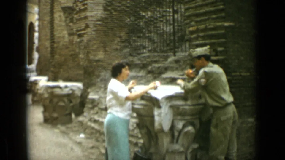 In 1962 Rome home-movie footage, a woman and a uniformed man stand by a stone table in a weathered brick courtyard.