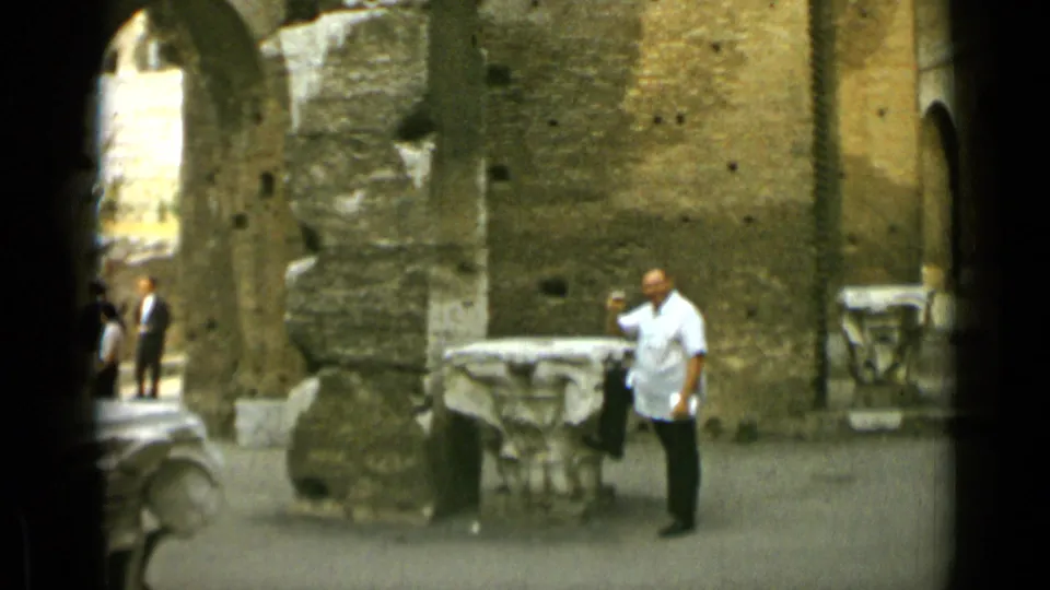 A man in a white shirt stands in the Rome courtyard near carved stone tables, with other visitors visible in the background.