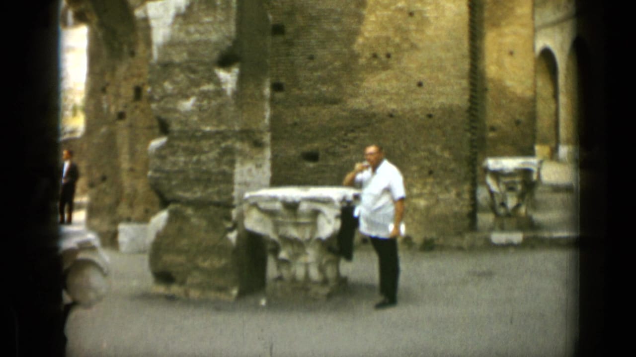 Man in a white shirt stands near stone ruins and a brick wall.