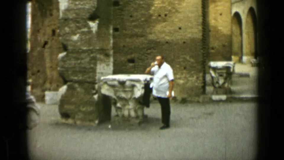 In the Rome travel scene, the man raises a drink while standing before ancient-looking stonework and weathered brick walls.