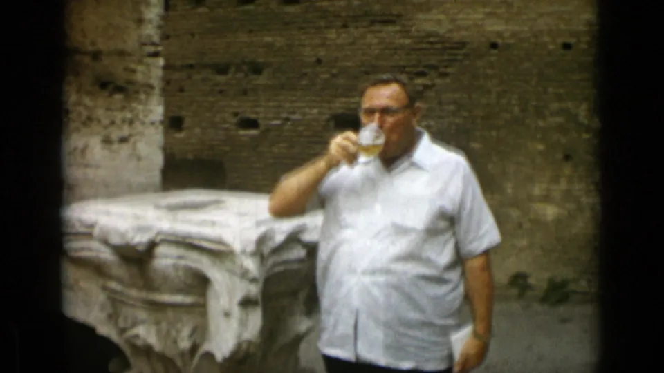 A closer 1962 Rome frame shows the man drinking from a beer glass beside a carved stone fragment and old brickwork.