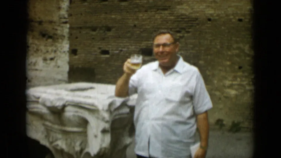 The man smiles and holds up his glass toward the camera during a 1962 Rome home-movie moment at a historic site.