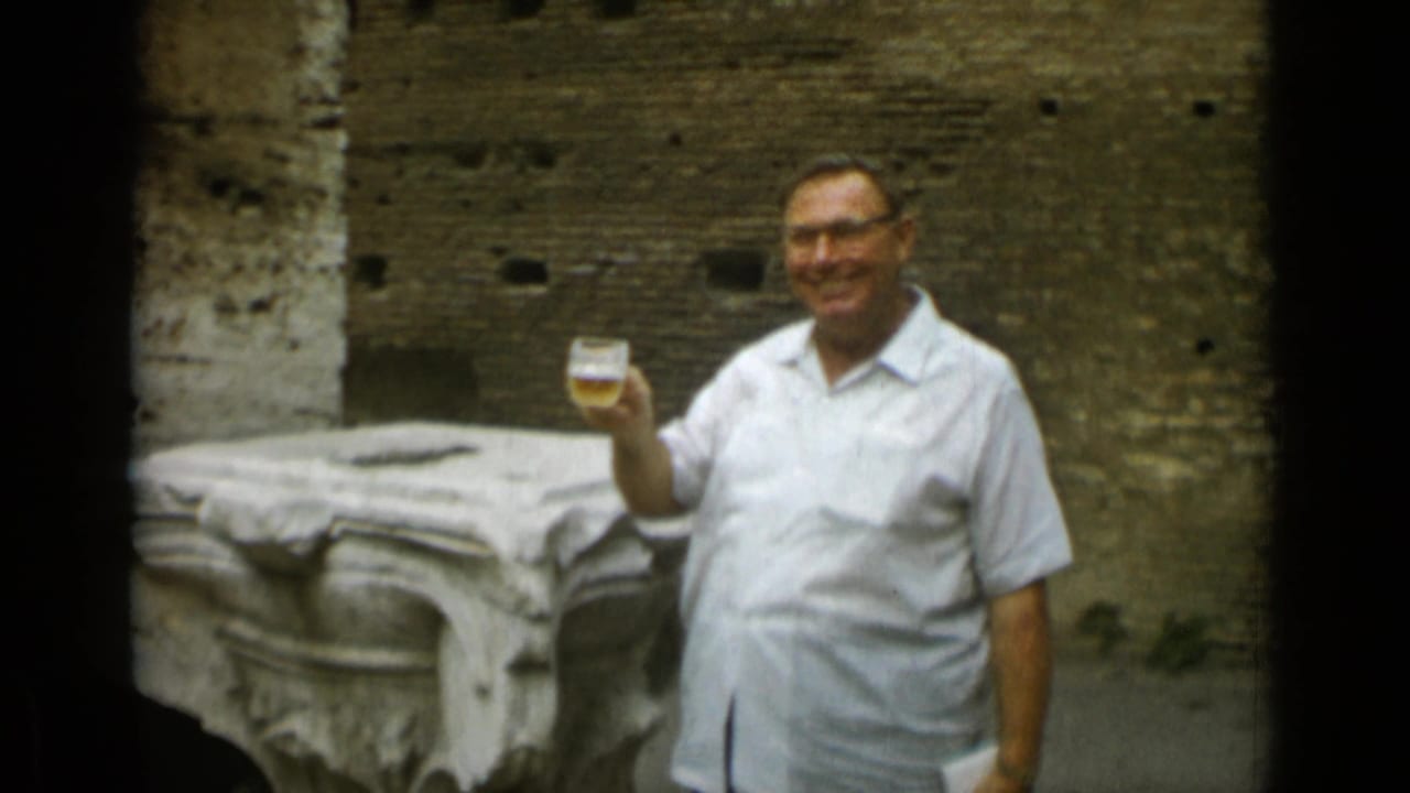 Man in a white shirt smiles and raises a beer glass beside old stone ruins.
