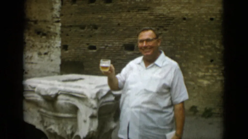 In Rome in 1962, a man in glasses and a white short-sleeve shirt smiles while holding up a glass of beer near ancient stonework.