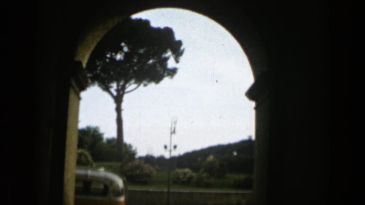 A visitor’s view through an arched opening in Rome shows an umbrella pine, roadway, lampposts, and part of a vintage orange and white bus.
