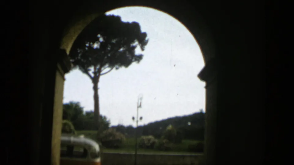A visitor’s view through an arched opening in Rome shows an umbrella pine, roadway, lampposts, and part of a vintage orange and white bus.