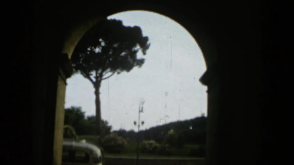 The camera looks from shadow through a rounded arch toward a sunlit Rome park scene with a tall pine, road, and partial orange and white bus.