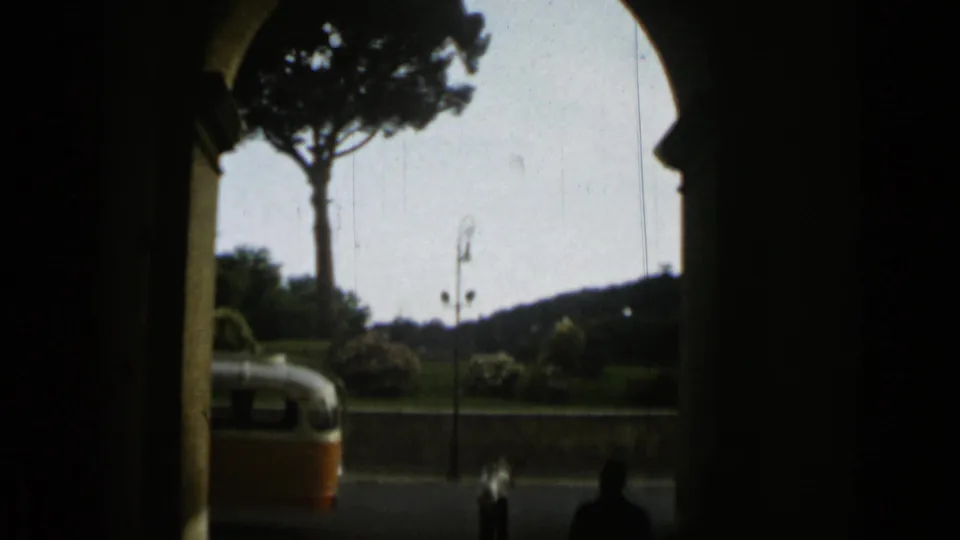 Through a dark arched opening in Rome, a tall tree, distant hillside, roadway, partial bus, and small silhouetted pedestrians are visible.