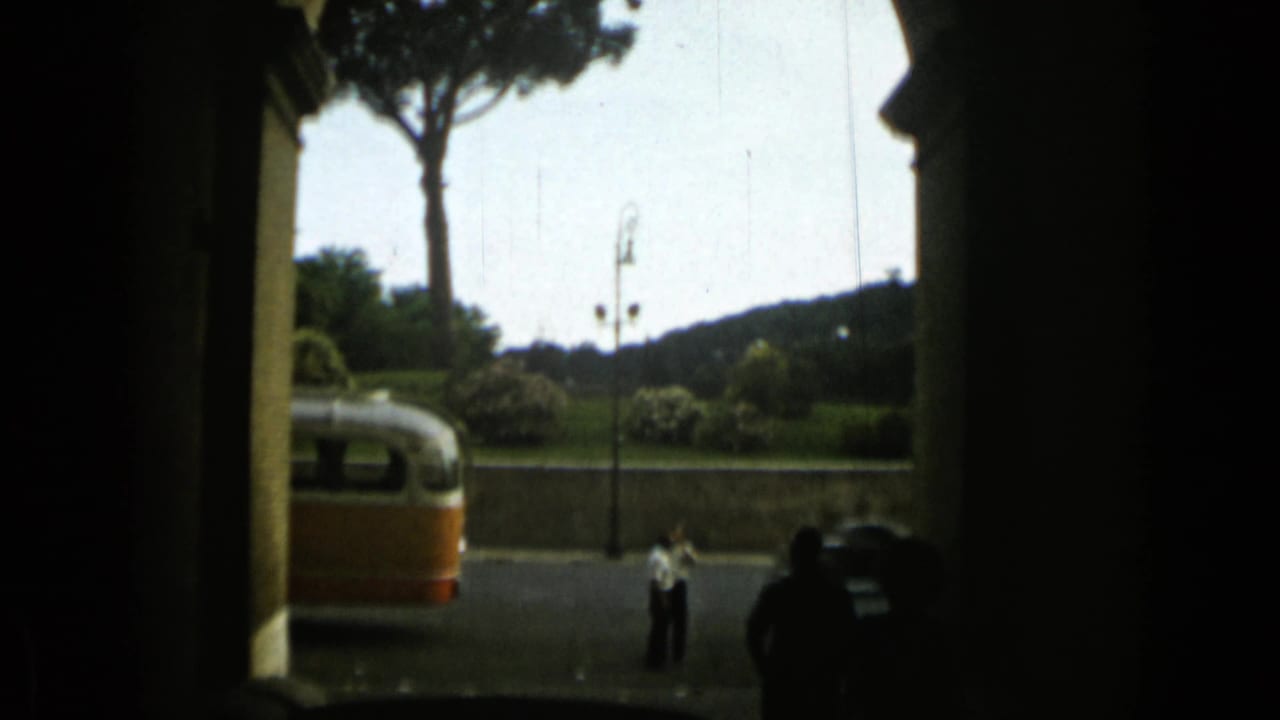 Street near the Colosseum seen through a shaded archway with a bus, cars, trees, and pedestrians.