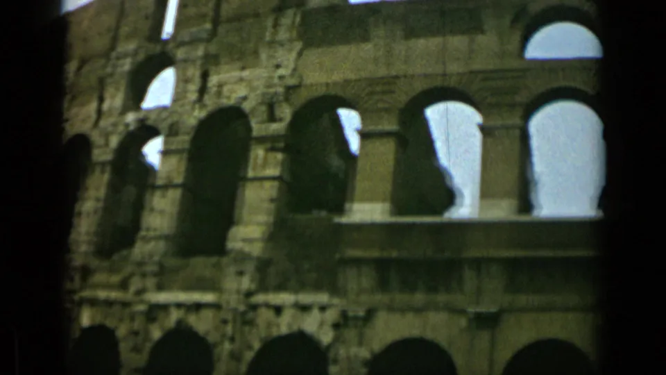 1962 Rome home-movie frame of the Colosseum wall, showing stacked arched openings, weathered stone, bright sky, and dark edge shadows.