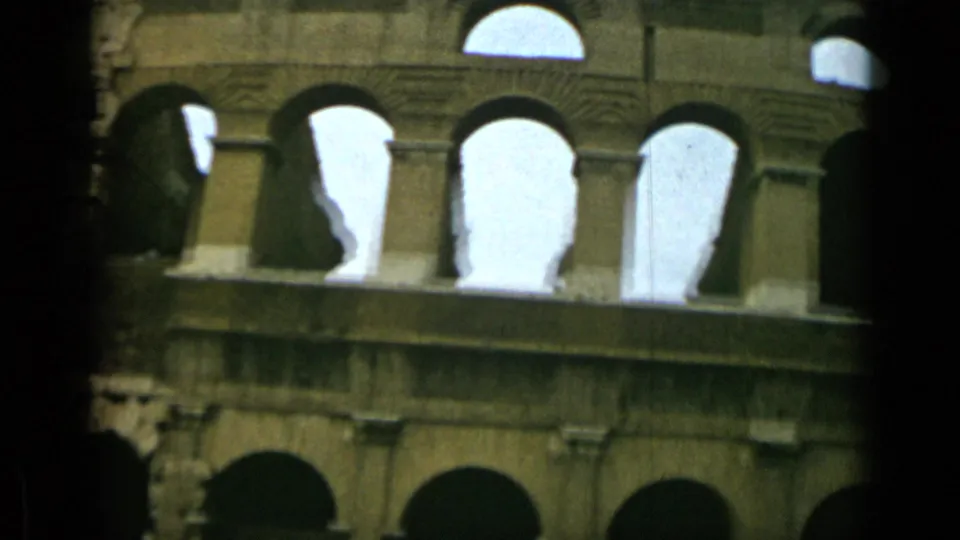 1962 Rome frame with the Colosseum facade filling most of the view, its upper arches silhouetted against bright sky in faded 8mm color.