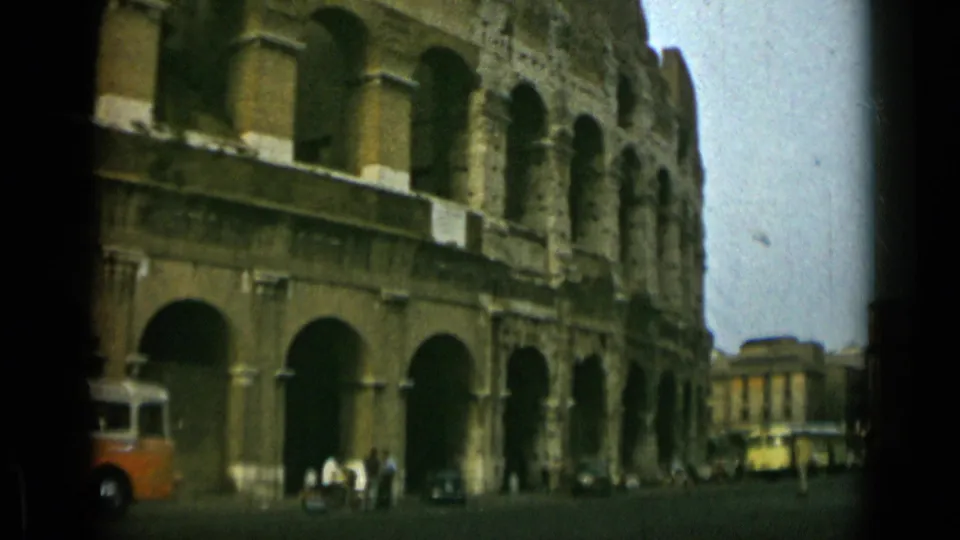 Wider 1962 Rome 8mm frame shows the Colosseum beside a street, with distant pedestrians and period vehicles below.