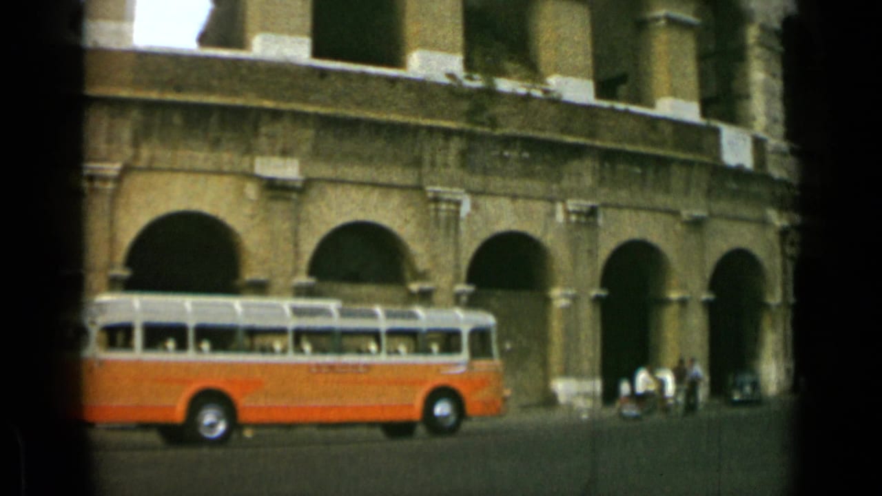 Orange-and-white bus stopped or moving beside the Colosseum arches.