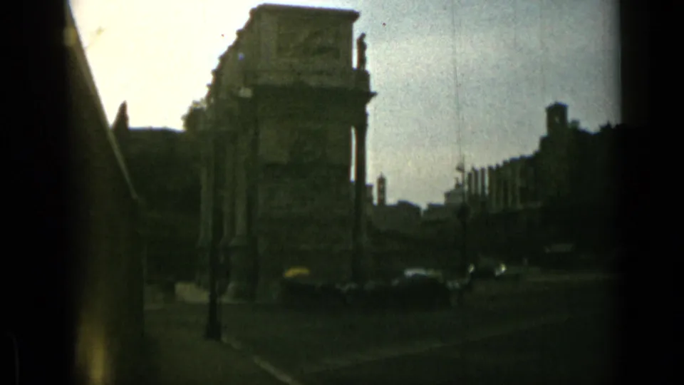 1962 Rome home-movie frame looking along a darker street near ancient ruins, with cars and silhouetted architectural forms.