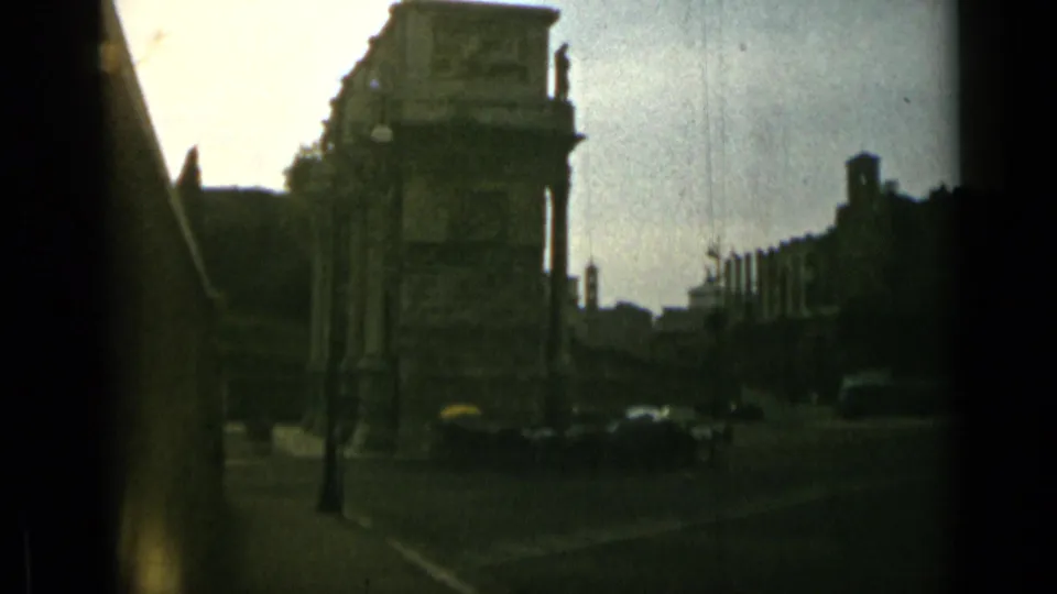 1962 Rome street view with a large stone archway, period cars, poles, and dark film-gate edges.