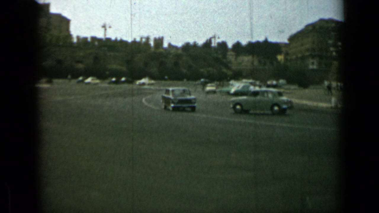 Rome street traffic in 1962, with small cars crossing an open road near trees and distant urban buildings.