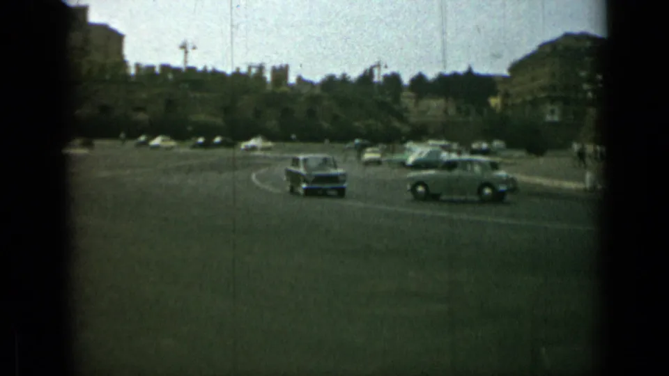 Rome street traffic in 1962, with small cars crossing an open road near trees and distant urban buildings.