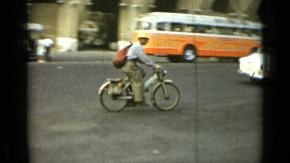 A bicyclist in a light shirt and hat rides through Rome traffic, with an orange-and-white bus behind him in 1962 footage.