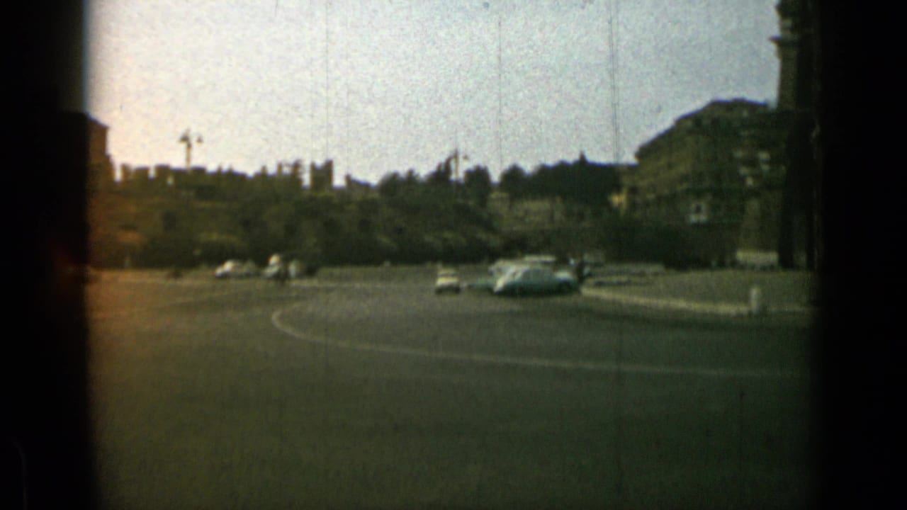 Wide road junction with several cars and distant buildings beyond a tree-lined edge.