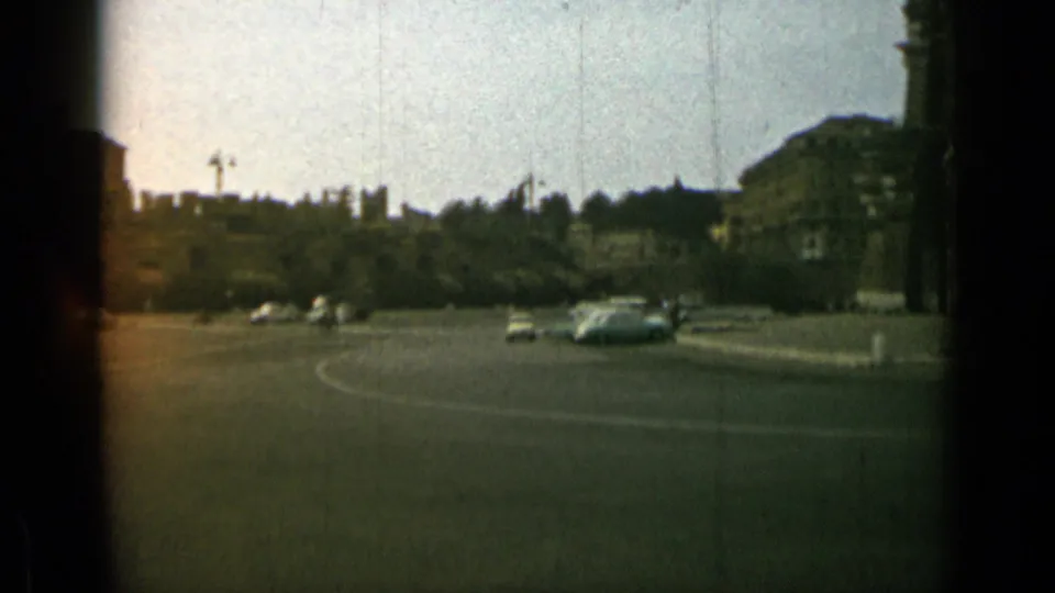 1962 Rome home-movie view of a broad roundabout with mid-century cars, trees, and distant urban buildings under pale daylight.