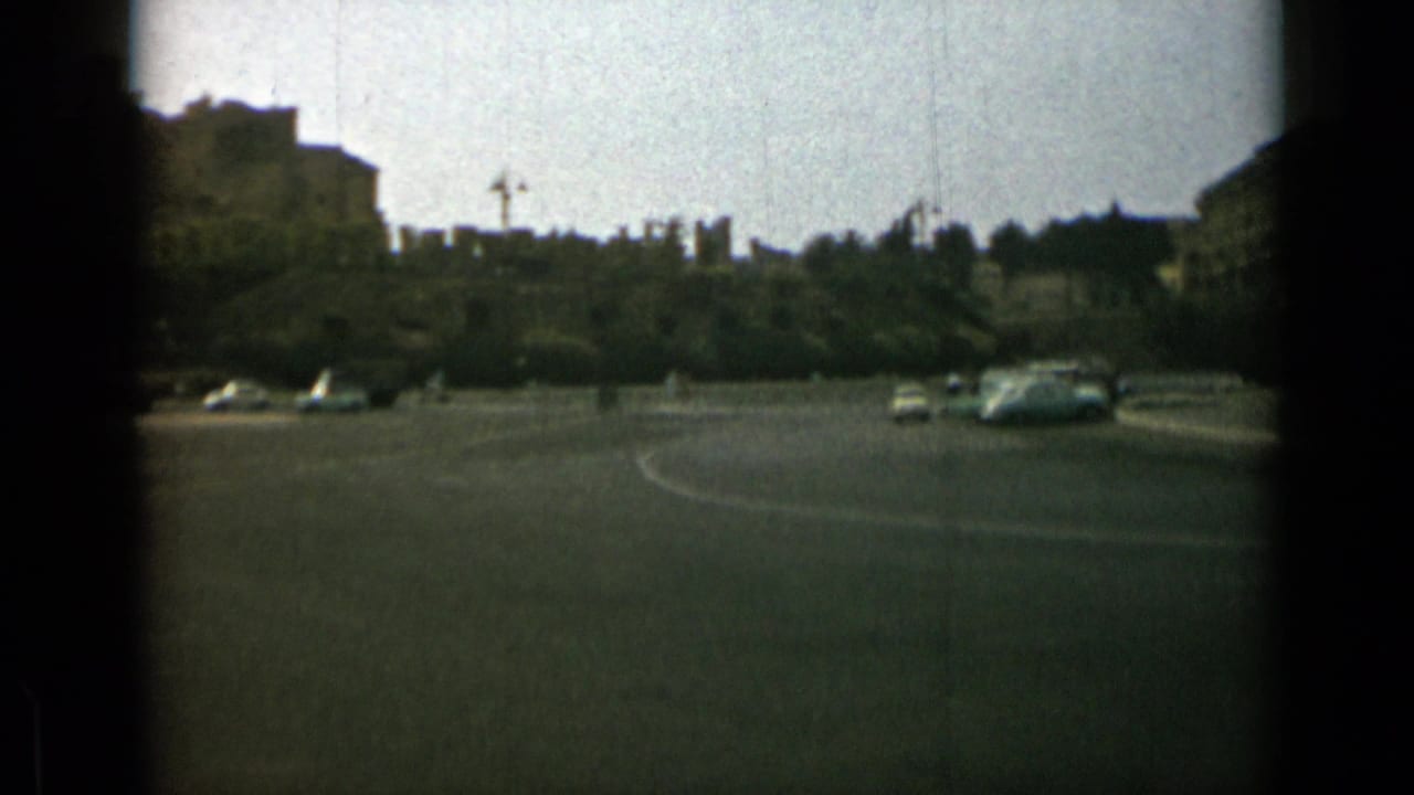 A 1962 Rome street view shows cars circulating across a wide roundabout, with trees and distant buildings softened by 8mm grain.
