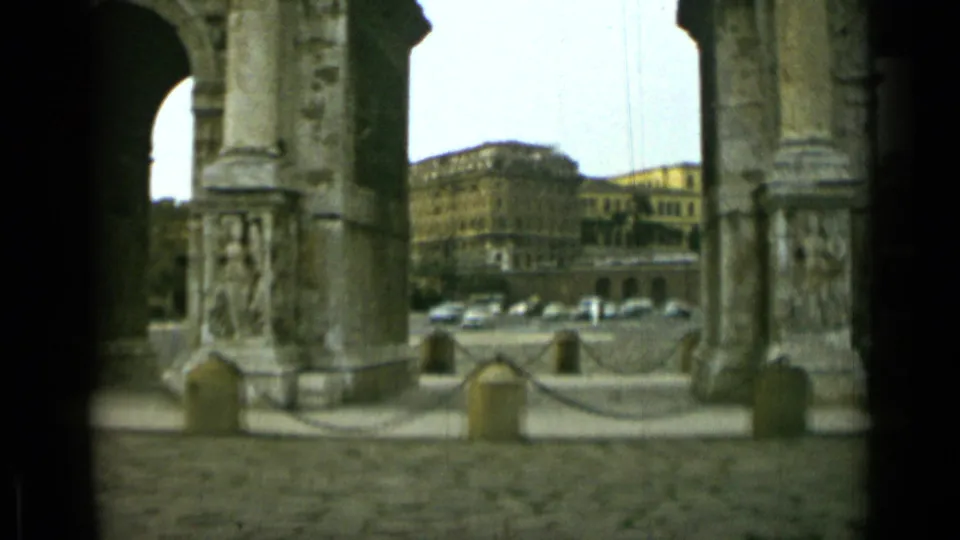 1962 Rome frame looking past a carved pier of the Arch of Constantine toward parked cars and mid-century buildings across the street.