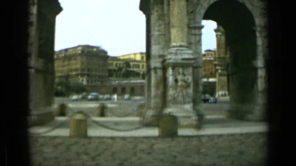 Rome 8mm frame from the Arch of Constantine, with a large arch opening, carved stone side, parked cars, and city buildings in clear daylight.