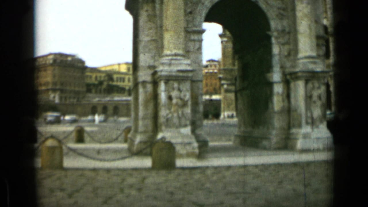 Stone archway and columns frame a cobblestone plaza with distant buildings.