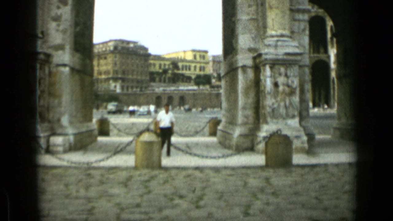 Man in a white shirt walks near chain barriers beneath large stone arches.