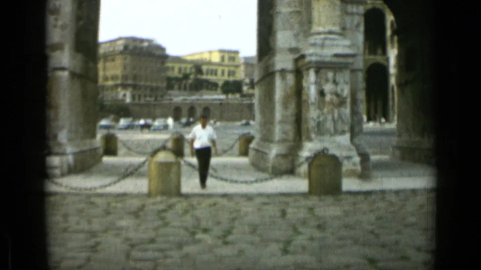 1962 Rome home-movie frame showing a man approaching through a stone archway with cobblestones and city buildings behind him.