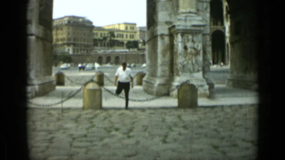 1962 Rome frame of a man moving through ancient arches, with cobblestones, chain barriers, and distant buildings visible.