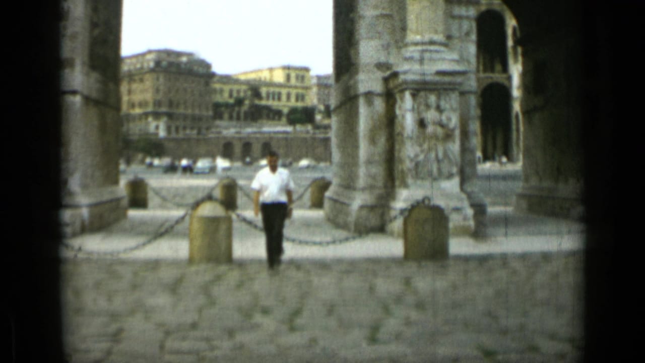 Man in a white shirt walks beneath ancient stone arches with city buildings beyond.