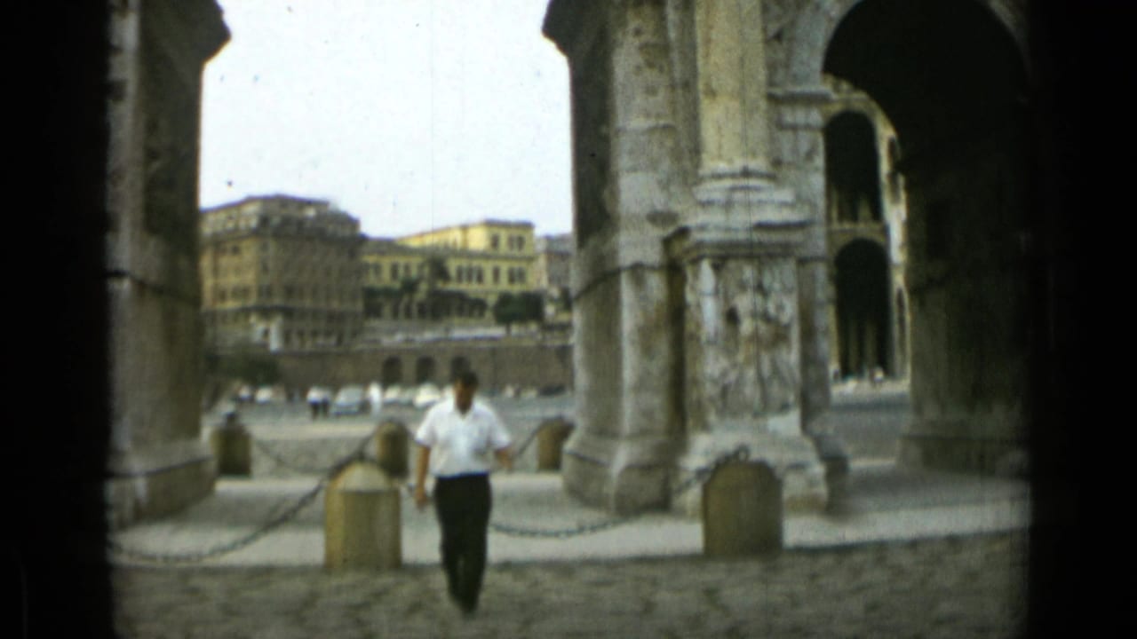 Man raises a hand while walking between stone arch supports and chain barriers.