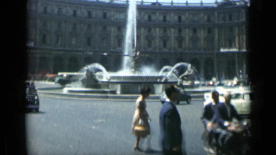 Rome street-square view with a large fountain, surrounding buildings, pedestrians, cars, and scooters captured in soft 8mm color.