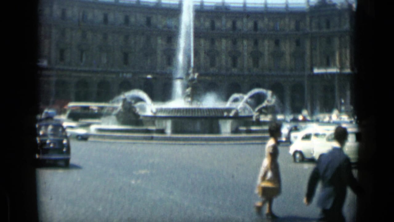 Traffic circle with fountain, cars, bus, and two pedestrians crossing in foreground.